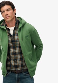 Hombre vistiendo una sudadera verde con cremallera sobre una camisa a cuadros azul y amarilla, con jeans, manos en los bolsillos, de pie contra un fondo blanco liso.