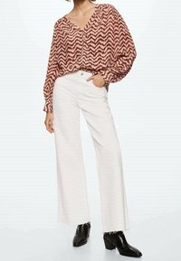 Donna che indossa una blusa a motivo chevron marrone, pantaloni bianchi a vita alta e gamba larga, e stivaletti neri, in piedi con una mano in tasca.