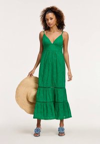 Roheline maksikleit sügava V-kaelusega, astmelise disainiga ja õrna tekstuuriga mustritega. Kombineeritud heinasügavusega kübaraga ja siniste volangiga sandaalidega.