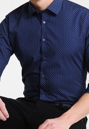 Man wearing a fitted navy blue shirt with a small white dot pattern and black pants, sitting with hands resting on thighs.