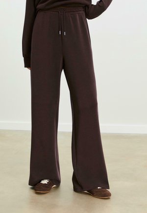 Personne portant un pantalon de survêtement large marron foncé avec taille élastique et cordon, associé à des baskets marron assorties sur un sol clair.