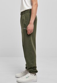 Olivengrønne joggebukser med elastisk liv, sidelommer og ribbestrikkede hems, sammen med hvite sneakers med grønne detaljer.