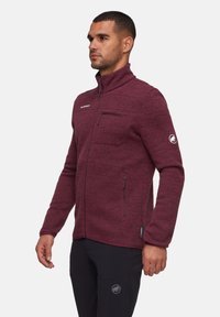 Giacca in fleece color bordeaux con colletto alto, tasca sul petto con zip e tasche laterali. Presenta un logo sul petto e una superficie texturizzata.