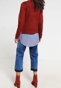 Sweter w kolorze rdzy z teksturowanym wykończeniem, nałożony na niebieską bluzkę w paski. W zestawie skrócone dżinsy i brązowe botki do kostek.