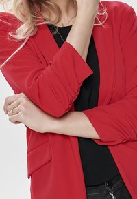 Blazer ligero rojo con mangas largas y un corte relajado. Presenta un bolsillo lateral y un sutil drapeado en las mangas. Diseño compacto.