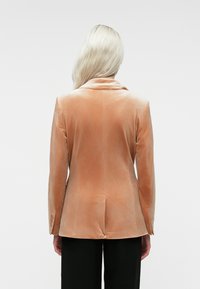 Une personne blonde se tenant de dos face à l'appareil, portant un blazer en velours couleur pêche et un pantalon noir, contre un fond uni.