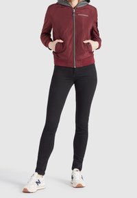 Vínová bomber bunda s kapucí, lehký materiál, zapínání na zip a boční kapsy. Černé úzké kalhoty a bílé tenisky s námořnickými akcenty.