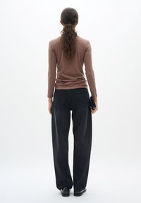 Femme aux cheveux bouclés en queue de cheval, portant un haut marron à manches longues, jean noir à jambes larges, chaussures noires, tenant une pochette noire, de dos.