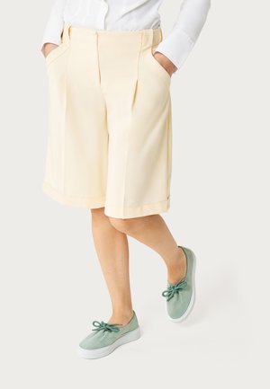 Persona vestida con pantalones cortos crema hasta la rodilla, camisa blanca de manga larga y zapatos verdes claros sin cordones con lazos en forma de lazo, manos en los bolsillos.