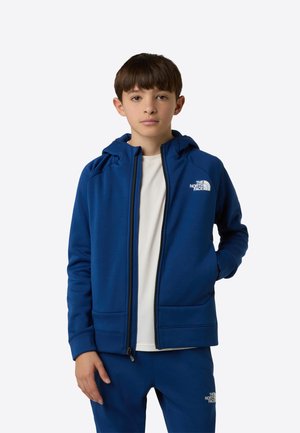 Sudadera azul con cremallera y capucha, con textura suave y puños acanalados. El logo está impreso en el lado izquierdo del pecho. Se lleva sobre una camisa blanca.