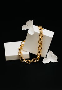 Guld kæde med store, indbyrdes sammenkoblede led, præsenteret på hvide geometriske blokke med hvide papir sommerfugle, sat mod en sort baggrund.