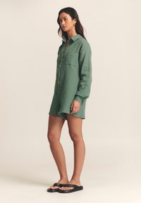 Zelena, teksturirana kratka srajčna obleka z ovratnikom, gumbi spredaj, enim prsnim žepom, dolgimi rokavi in sproščenim krojem, kombinirana s črnimi sandali.
