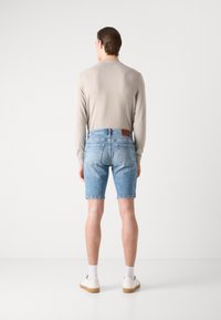 Camisa de manga longa bege claro combinada com calções de ganga azul claro, apresentando um retalho de pele castanha na parte traseira da cintura. Ténis brancos e meias.