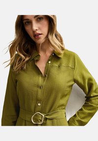 Camicia a maniche lunghe verde oliva con chiusura a bottoni, tessuto testurizzato e cintura in vita con una fibbia dorata di forma irregolare.