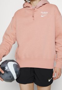 Persona che indossa una felpa rosa del Paris Saint-Germain e pantaloni corti neri della Nike, mentre tiene in mano un pallone da calcio Nike bianco e nero.