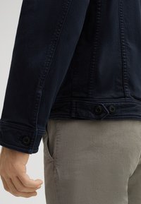 Giacca di jeans blu navy con polsini e orlo con bottoni, caratterizzata da una linea dritta e un tessuto testurizzato. Pantaloni beige visibili sotto.