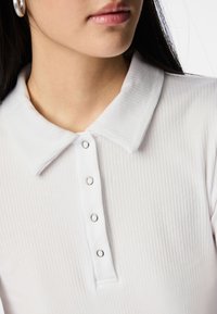 Bílý žeberný top s límečkem, pět stříbrných patentek a měkká textura. Důraz je kladen na detaily límce a knoflíků.