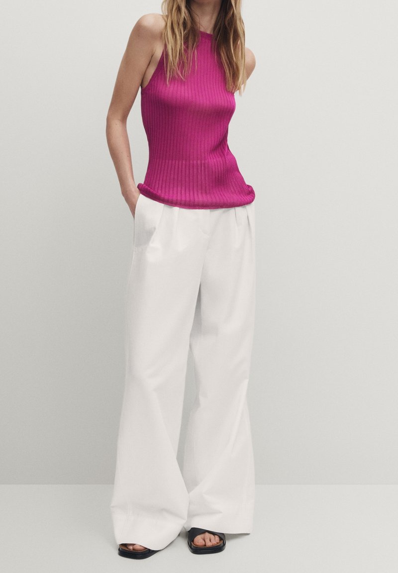 Femme portant un haut magenta côtelé sans manches, un pantalon large blanc ample et des sandales noires à bouts ouverts, debout les mains dans les poches.