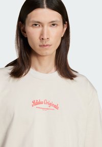 Smėlinis medvilnės marškinėliai su apvaliu kaklu ir trumpomis rankovėmis. Priekyje yra „Adidas Originals“ žodis rožinės spalvos skaitmeninėse raidėse.