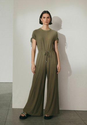 Olivengrøn jumpsuit med korte ærmer, snor i taljen og to forlommer; bred ben-design og letvægtsstof. Sorte sandaler.