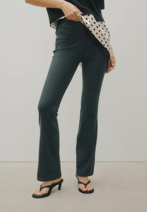 Persona che indossa pantaloni neri aderenti, sandali neri con tacco alto e una maglietta nera con uno strato interno beige a pois, in piedi su uno sfondo uniforme.