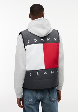 Bărbat purtând hanorac gri, vestă matlasată închisă la culoare cu logo mare Tommy Jeans roșu, alb și negru, și blugi albaștri, văzut din spate.