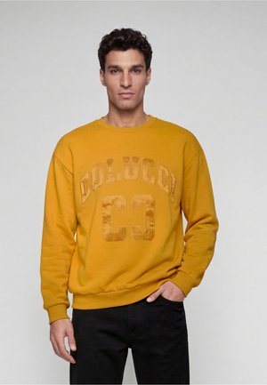 Homme portant un sweat jaune moutarde avec le texte "COLUCCI CC", debout devant un fond gris clair uni avec une main dans la poche.
