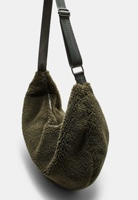 Olijfgroene schoudertas met een zachte textuur en een gebogen vorm, voorzien van een ritssluiting en een verstelbare schouderband van duurzaam materiaal.