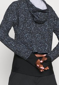 Zkrácený černý sportovní top s modrými skvrnami, má kapuci a dlouhé rukávy, vyrobený z tkaniny, která odvádí vlhkost.