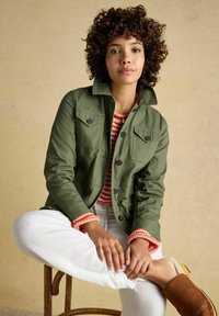 Jauna moteris su garbanotais plaukais, dėvinti žalią striukę, raudonai baltą dryžuotą megztinį, baltas kelnes ir rudus slip-on batus, sėdi sukryžiavusi kojas.