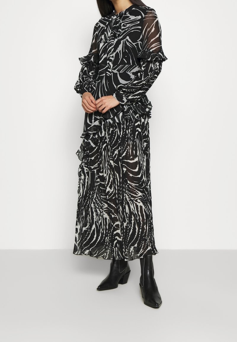 Robe maxi transparente noir et blanc à motif zèbre, manches à volants et détail noué au col, associée à des bottes noires pointues.