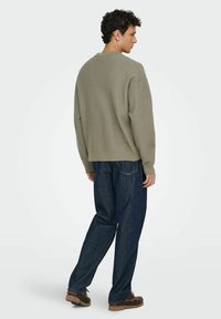 Maglione verde oliva con una vestibilità rilassata, colletto e polsini a coste, abbinato a jeans in denim blu scuro e scarpe marroni, visto da dietro.