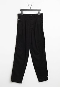 Pantalones de algodón negros con un corte holgado, que presentan pinzas frontales, un cierre de botón y bolsillos a los lados. Colgados en una percha negra.