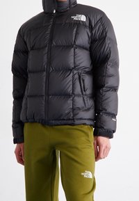 Man i svart vadderad North Face-jacka och olivgröna North Face-sweatpants, som står mot en enhetlig bakgrund.