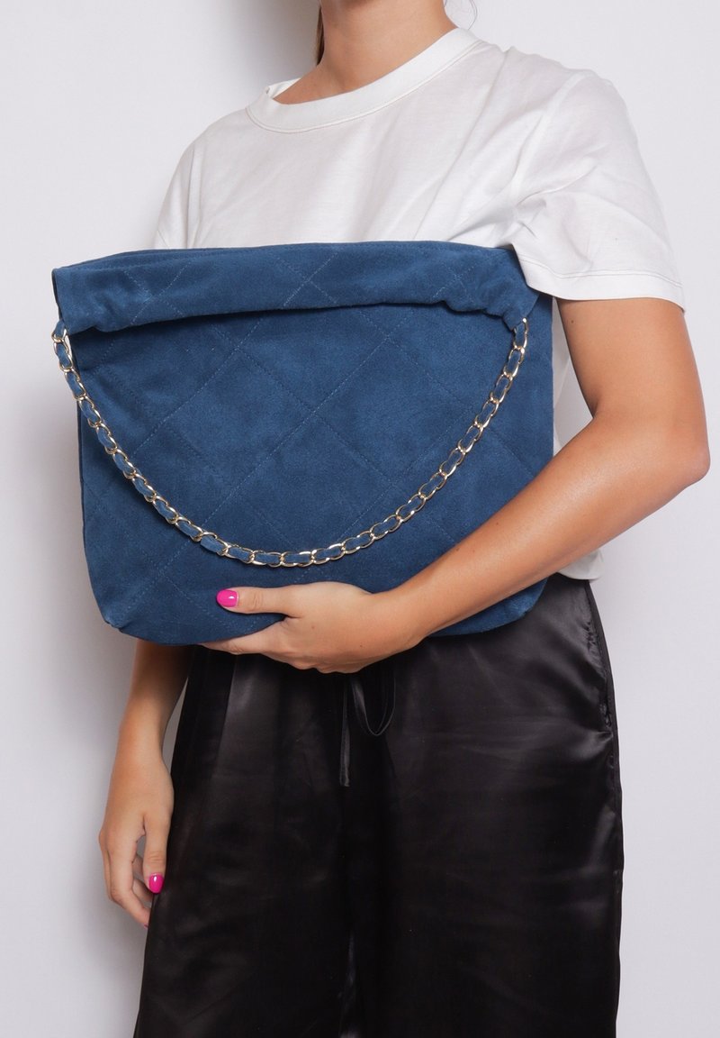 Borsa in suede blu trapuntato con un cappuccio pieghevole, dotata di una tracolla in argento. La borsa è tenuta di fronte a una persona che indossa una camicia bianca.
