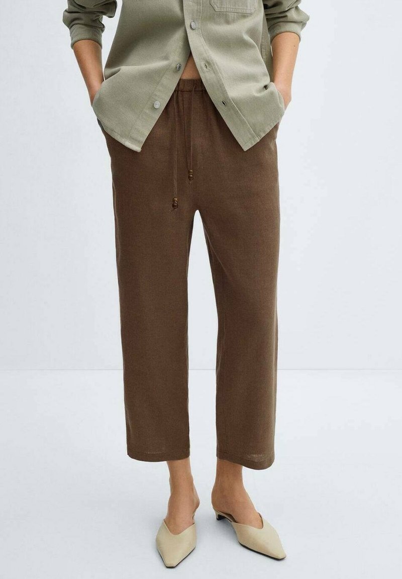Pantalon marron ample avec taille à cordon, ourlets retroussés et texture douce, associé à une chemise boutonnée vert clair et des chaussures beige.
