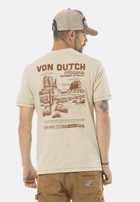 Camiseta de algodón beige con un gráfico marrón de monumentos del desierto y cactus. También incluye detalles de texto "VON DUTCH" y "Arizona".