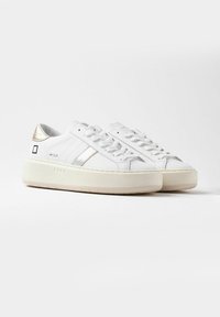 Zapatillas blancas con parte superior de cuero suave, rayas plateadas y acento en el talón de color dorado metálico. Suela de goma blanca gruesa con textura estriada.