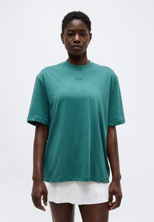 Mujer con el pelo corto y rizado, vistiendo una camiseta suelta de color verde azulado sobre una falda blanca, de pie frente a un fondo claro y liso.