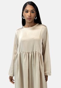 Kremfarvet satin kjole med lange ærmer, rund hals og indsnævret talje. Stoffet har en glat tekstur og let glans.