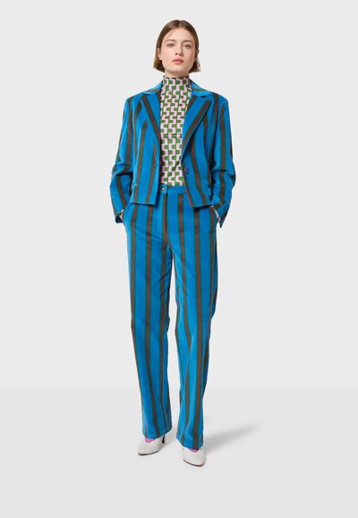 Plavi i smeđi prugasti odijelo s prilagođenim sakoom i ravnim hlačama. Slojevito s zelenim turtleneckom s geometrijskim uzorkom. Bijele štikle.