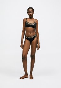 Frau mit geflochtenem Haar, die einen schwarzen Bikini mit beigefarbenen Wellenmustern und braunen flachen Sandalen trägt, steht vor einem schlichten weißen Hintergrund.