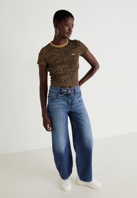 Žena oblečená v upnutém tričku s leopardím potiskem, širokých modrých džínách s uvázaným opaskem, bílých teniskách a velkých kruhových náušnicích, pózující před jednoduchou zdí.