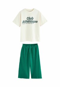 Camiseta de algodón color crema con texto verde oscuro que dice "Club Athlétique Track And Field Dept." combinada con pantalones cortos de pierna ancha verdes.