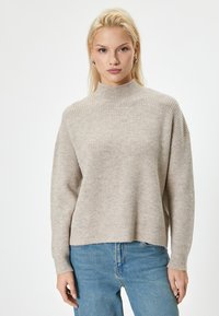 Beżowy sweter prążkowany z wysokim kołnierzem, szerokimi rękawami i luźnym krojem, w połączeniu z jasnoniebieskimi dżinsami. Miękka tekstura i swobodny styl.