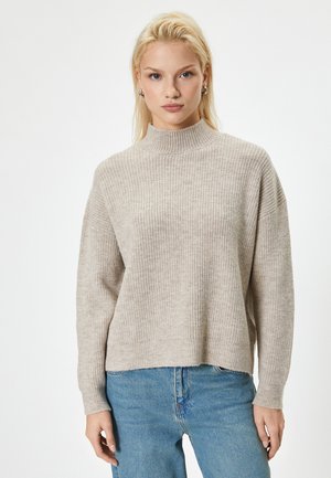 Pull beige côtelé avec un col haut, des manches larges et une coupe décontractée, associé à un jean en denim bleu clair. Texture douce et design décontracté.