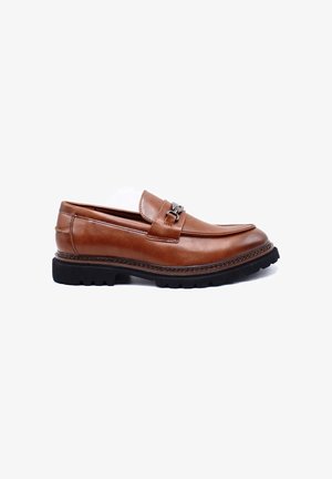 Mocassins en cuir marron avec un bout rond, une sangle décorative et des éléments en argent, reposant sur une semelle en caoutchouc noir texturé.