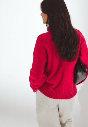 Kvinde med langt mørkt hår iført en knaldrød sweater, lyse bukser og holder en sort håndtaske bag ryggen.