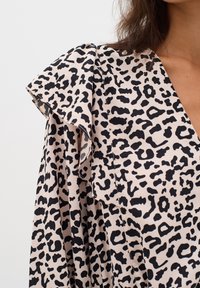 Leopardmönstrad klänning med svarta fläckar på en beige bakgrund, som har en draperad axeldesign och en djup V-ringning. Slät tygtextur.