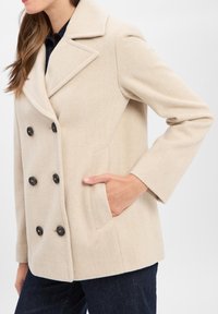 Frau trägt einen beigen doppelt geknöpften Wollmantel mit schwarzen Knöpfen, die Hand in der Tasche, kombiniert mit dunkelblauer Jeans.